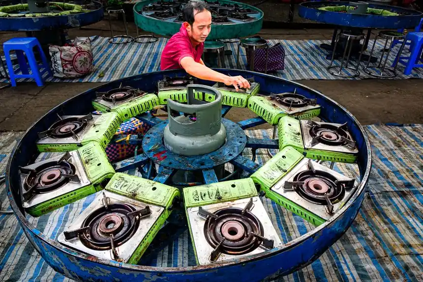 A man arranges stove burners around a central gas cylinder outdoors.
